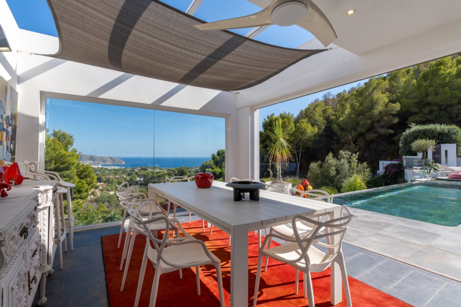 Comedor exterior acristalado con vistas panorámicas al mar, mesa blanca de diseño, ventilador de techo y acceso directo a la piscina infinita rodeada de vegetación.