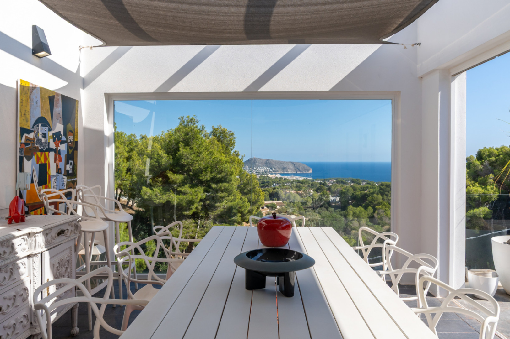 Comedor exterior con mesa blanca y sillas de diseño bajo un toldo vela, destacando las vistas panorámicas al mar y a la costa de Moraira a través de ventanales.