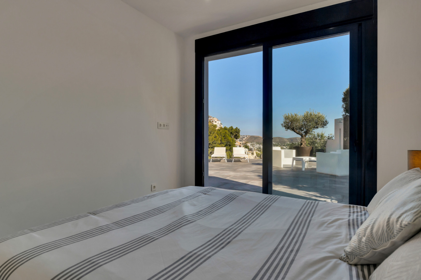 Dormitorio con acceso directo a la terraza mediante grandes ventanales. Ofrece vistas despejadas al área de descanso exterior, un olivo ornamental y las montañas.