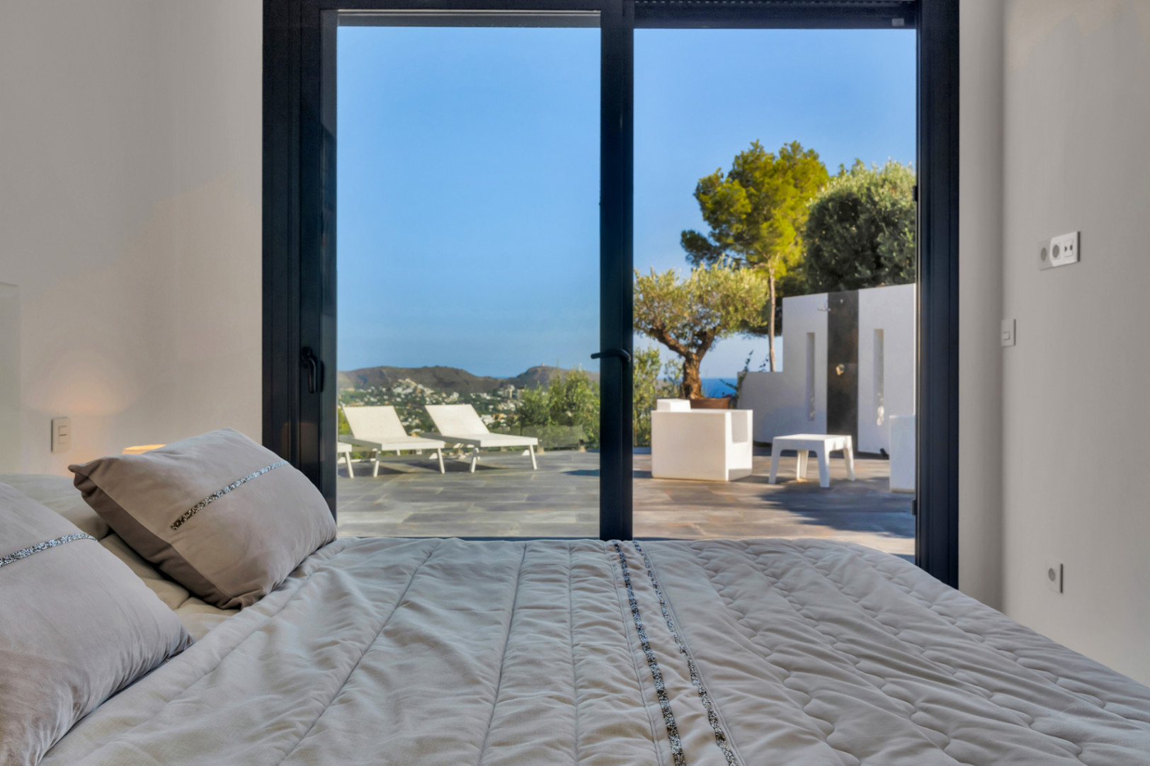 Dormitorio con acceso directo a la terraza mediante ventanales correderos, ofreciendo vistas al mar, mobiliario exterior blanco y zona de ducha de verano.