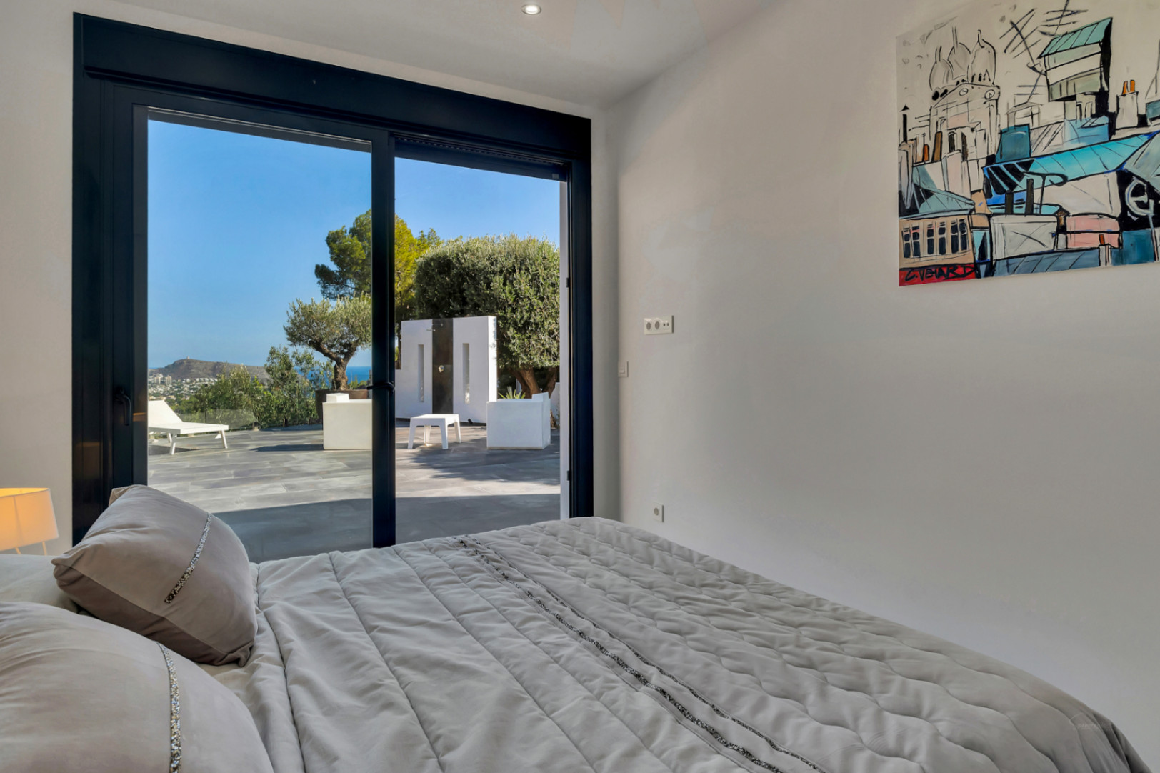 Dormitorio con acceso directo a la terraza mediante ventanales de suelo a techo, ofreciendo vistas al mar y al jardín mediterráneo desde la cama.