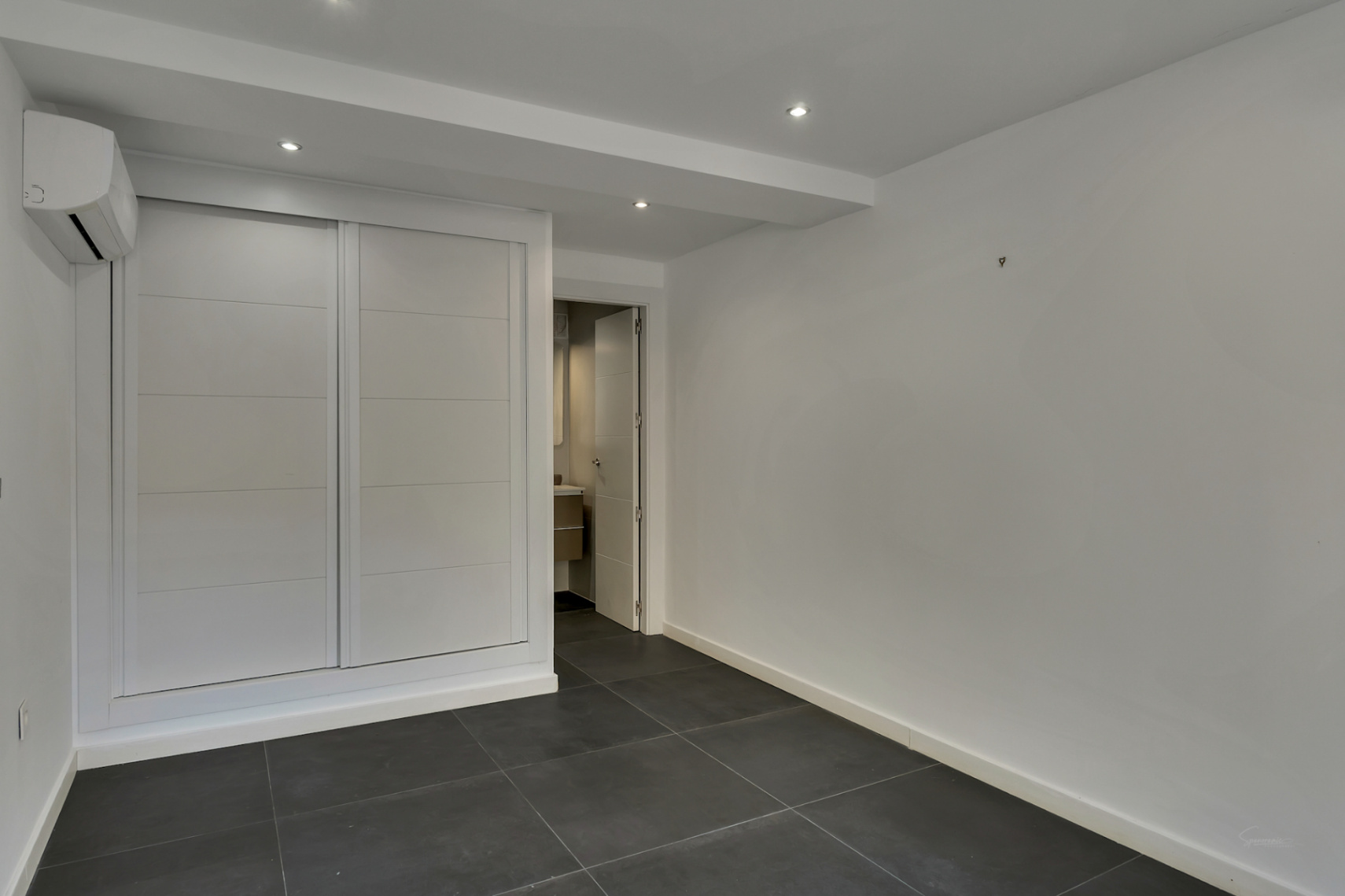 Dormitorio con armario empotrado de puertas correderas blancas, suelo de gres porcelánico oscuro, aire acondicionado y acceso directo a baño en suite.