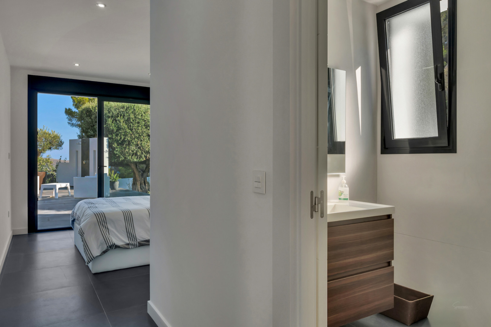 Dormitorio en suite con baño integrado y acceso directo a la terraza. Presenta suelos cerámicos oscuros, mueble de madera y vistas al jardín mediterráneo con olivo.