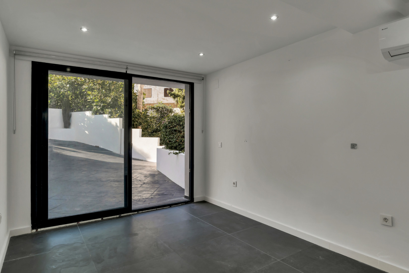 Dormitorio o despacho con suelos de gres oscuro, iluminación LED empotrada y gran ventanal corredero con acceso directo a la zona exterior pavimentada.