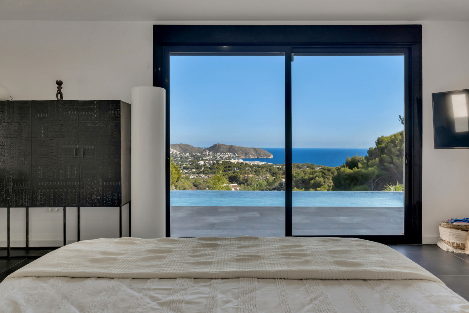 Dormitorio principal con vistas panorámicas al mar y la costa de Moraira a través de grandes ventanales. Incluye acceso directo a la terraza y la piscina infinita.