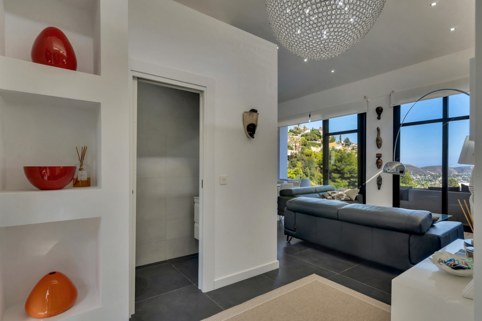 Entrada luminosa con hornacinas decorativas integradas, suelos de gres oscuro y vistas al salón principal con grandes ventanales hacia el paisaje mediterráneo.