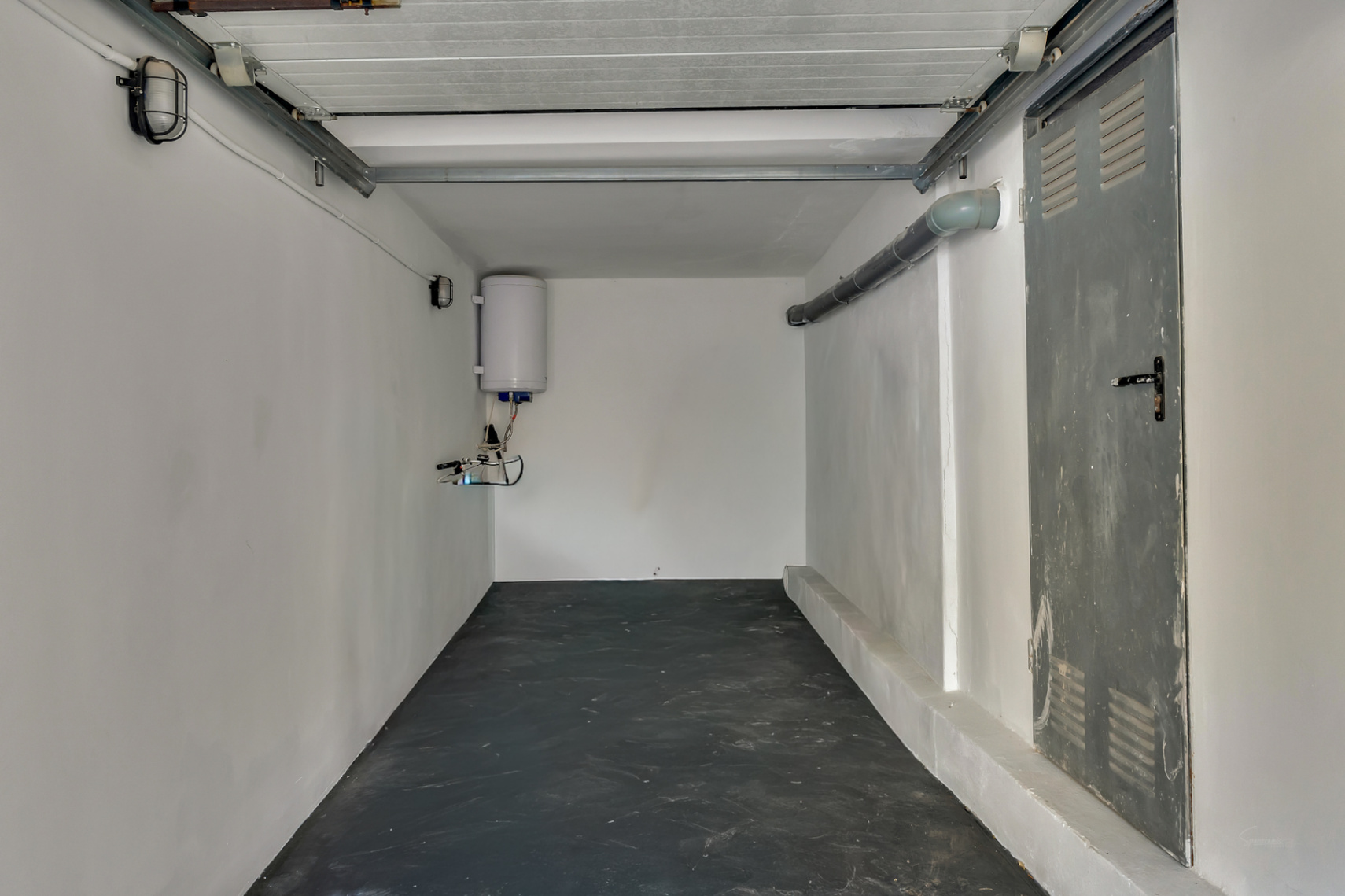 Garaje funcional con suelo oscuro, paredes blancas, termo eléctrico y puerta metálica lateral, ideal para almacenamiento o vehículos pequeños.