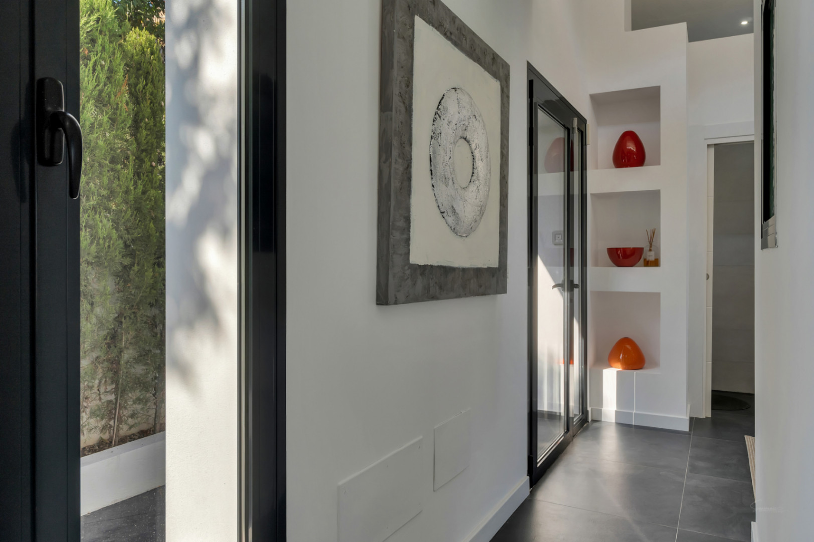 Pasillo de diseño con suelo porcelánico oscuro, hornacinas decorativas integradas y ventanales de carpintería negra que aportan luz natural y vistas al jardín.