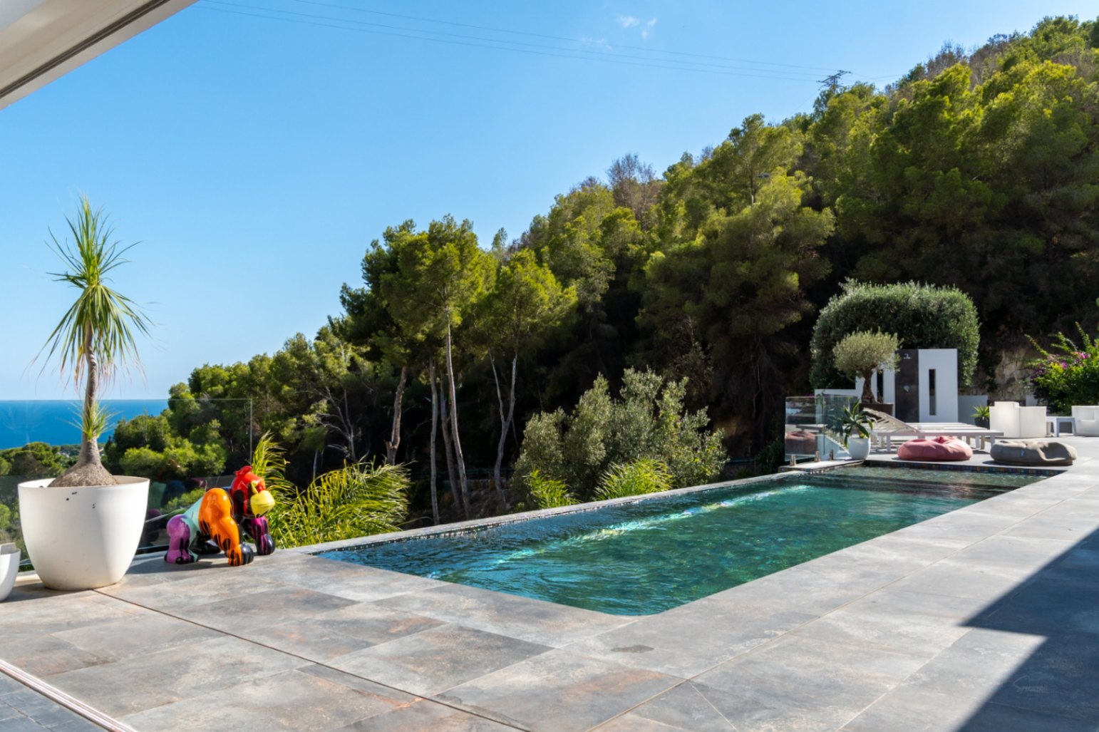 Piscina desbordante con vistas al mar y la montaña, integrada en una terraza de gres porcelánico gris con una escultura decorativa y entorno forestal.