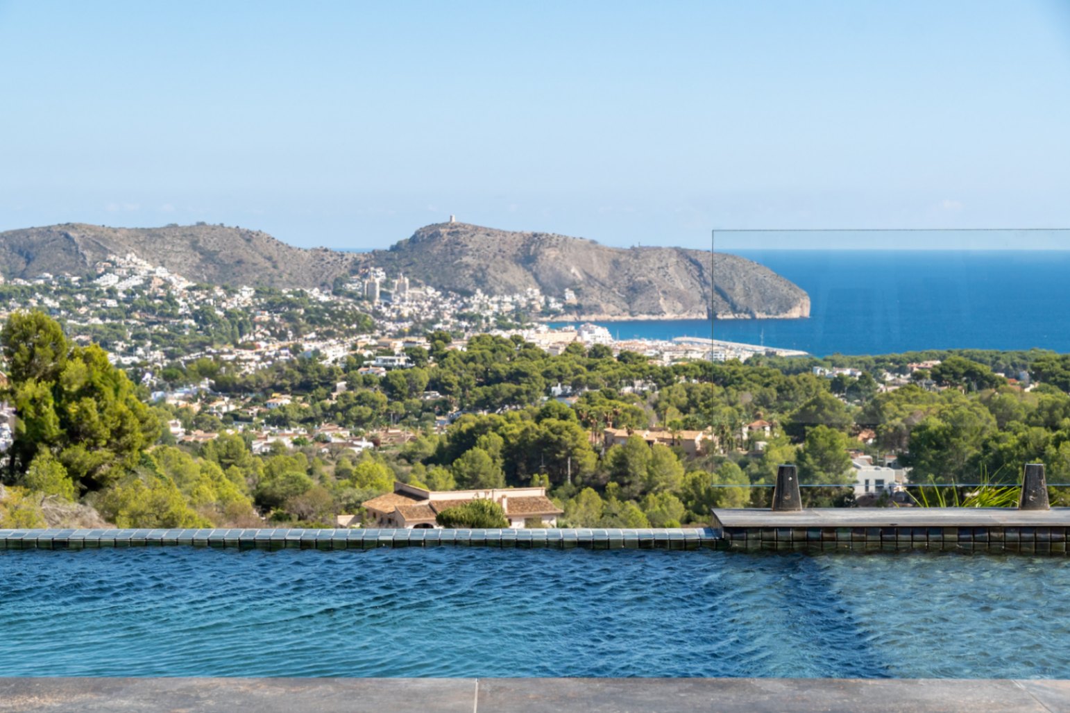 Piscina infinita con borde de gresite azul y barandilla de cristal, ofreciendo vistas panorámicas al mar Mediterráneo, el Cap d'Or y el entorno verde de Moraira.
