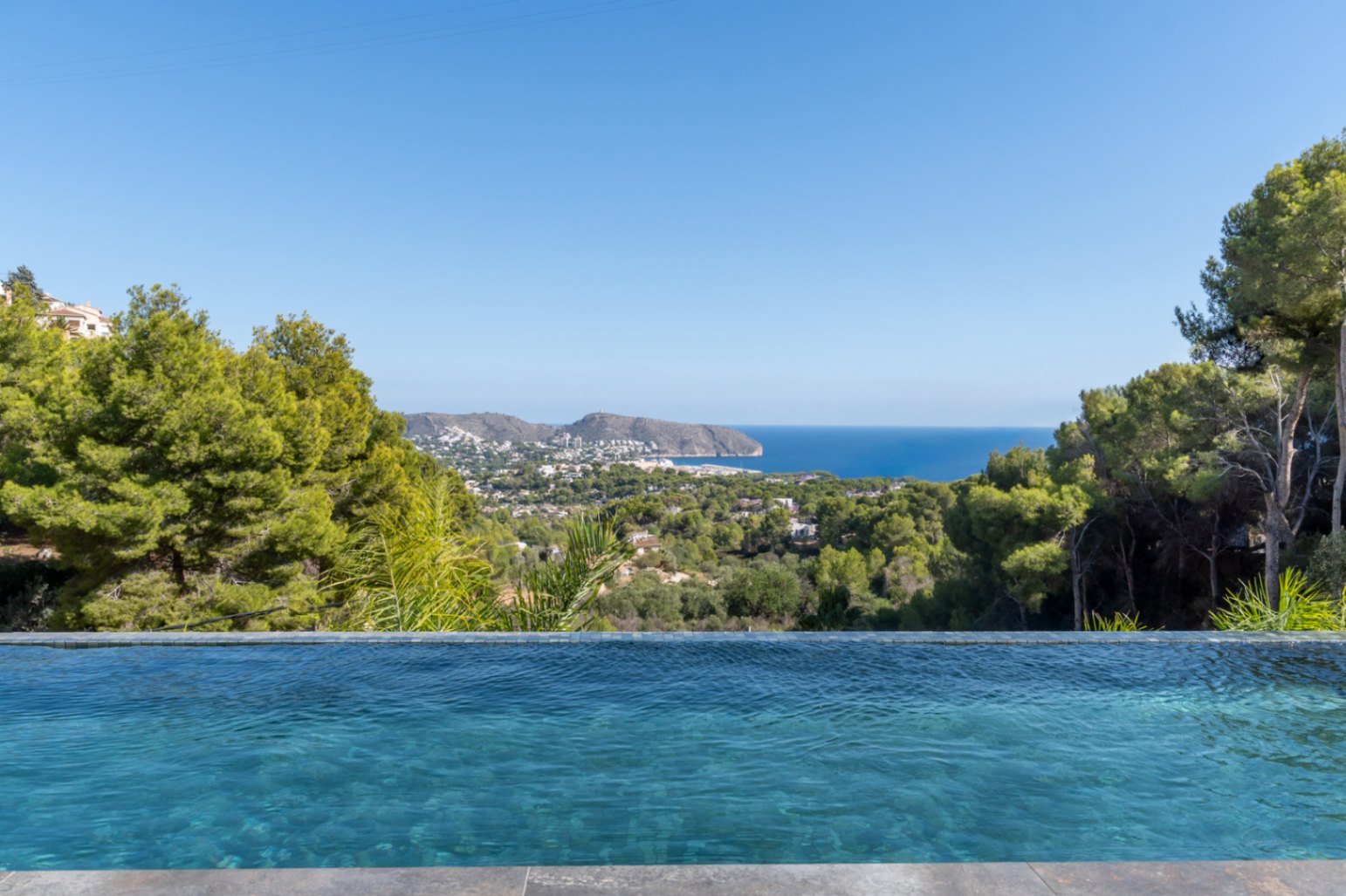 Piscina infinita con revestimiento de mosaico azul y vistas panorámicas al mar Mediterráneo, la costa de Moraira y el entorno boscoso circundante.