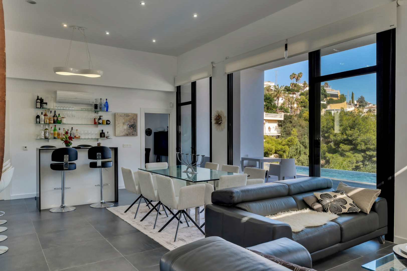 Salón-comedor de planta abierta con techos altos, zona de bar integrada y grandes ventanales que ofrecen vistas directas a la piscina y al entorno mediterráneo.