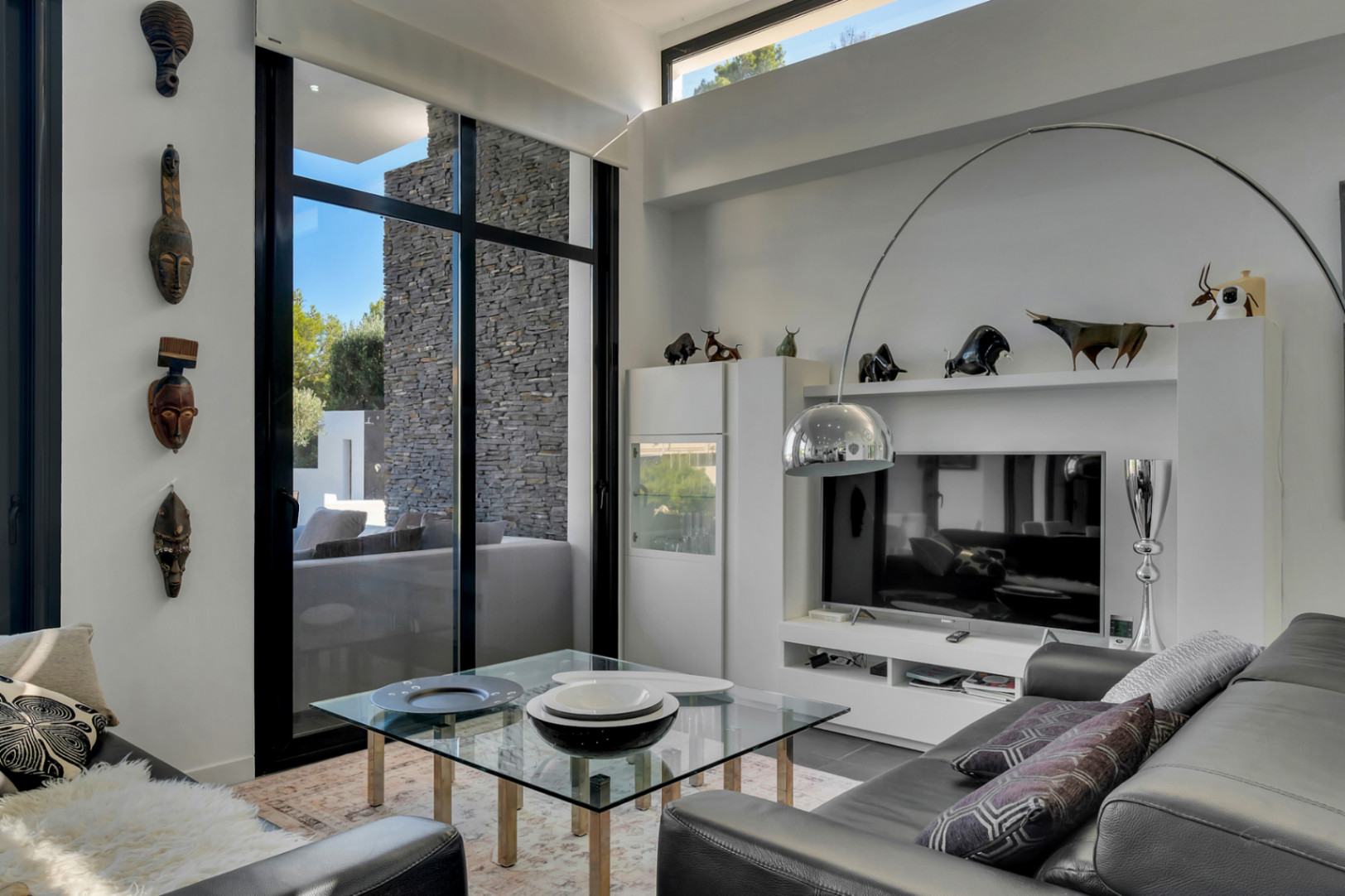 Salón de diseño con techos de gran altura, ventanales negros y decoración étnica. Destaca la lámpara de arco cromada y el acceso visual al muro de piedra natural exterior.