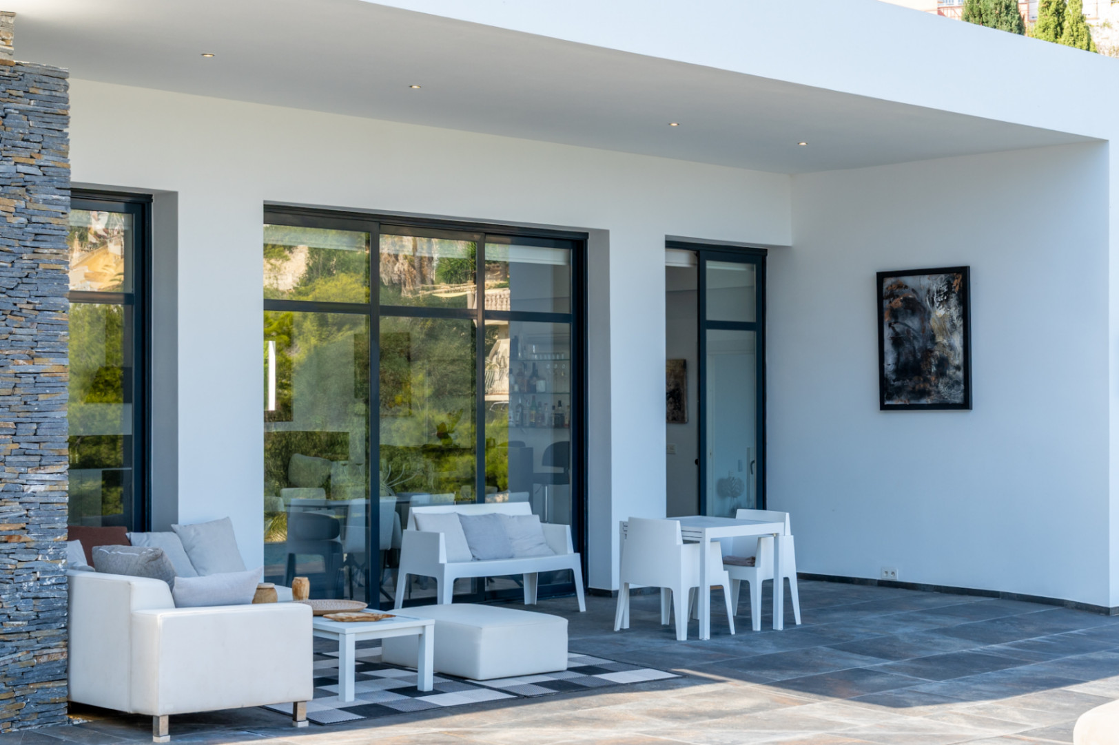 Terraza cubierta con suelos de gres oscuro, pilares de piedra natural y mobiliario minimalista blanco frente a grandes ventanales de carpintería antracita.