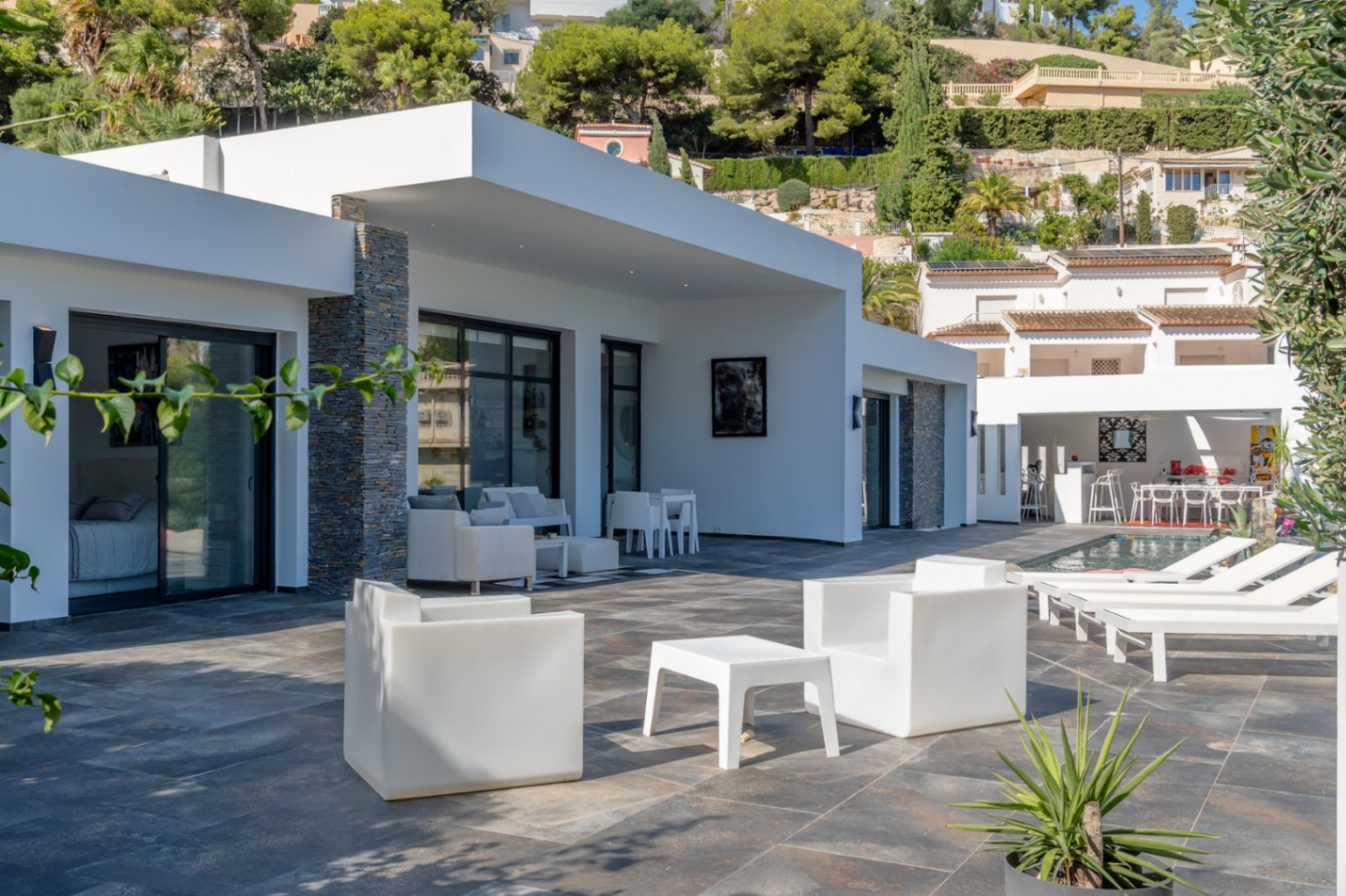 Terraza principal con pavimento de piedra oscura, mobiliario de diseño blanco y fachada con detalles en piedra natural junto a la zona de piscina.