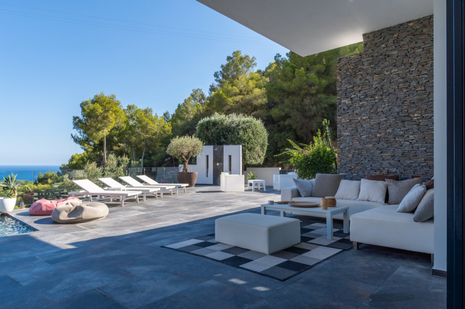 Terraza principal con zona de estar, sofás modulares blancos y pared de piedra natural. Al fondo, solárium con tumbonas y vistas panorámicas al mar Mediterráneo.