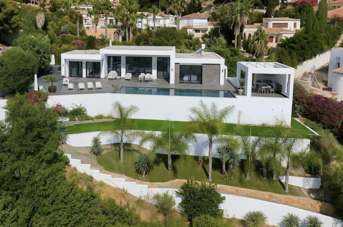 Vista exterior de la villa con arquitectura moderna, terraza principal, piscina infinita y jardín mediterráneo escalonado con palmeras.