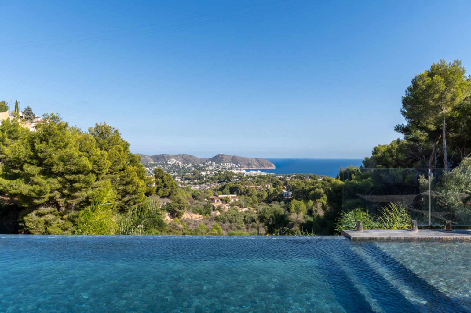 Vista panorámica desde la piscina infinita hacia el mar Mediterráneo y las montañas, rodeada de vegetación autóctona y protegida por una barandilla de cristal.