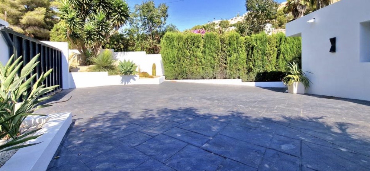 Zona de aparcamiento con pavimento de hormigón impreso oscuro, rodeada de muros blancos, setos de ciprés y vegetación mediterránea con yucas y agaves.