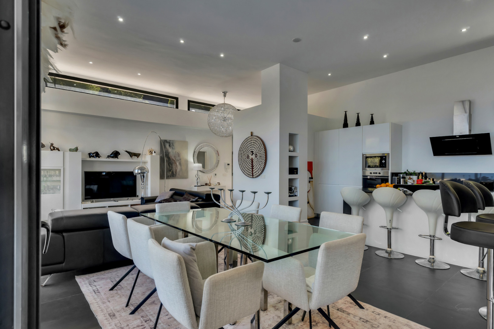 Zona de día diáfana con comedor de cristal, cocina de diseño con barra y techos altos que integran ventanales superiores y focos empotrados.