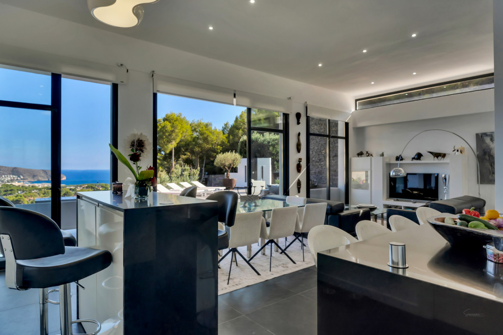 Zona de día diáfana con techos altos, grandes ventanales con vistas al mar y cocina integrada con barra negra conectada al salón-comedor.