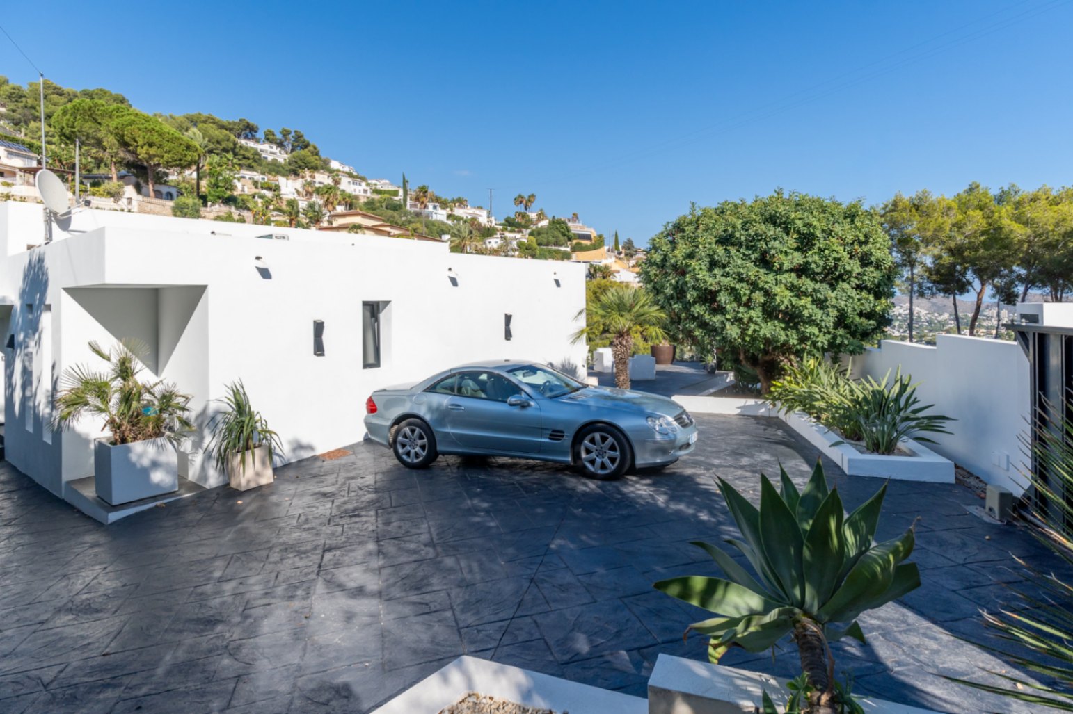 Zona de entrada y parking con pavimento de hormigón impreso oscuro, fachada minimalista blanca y jardineras integradas con vegetación mediterránea.