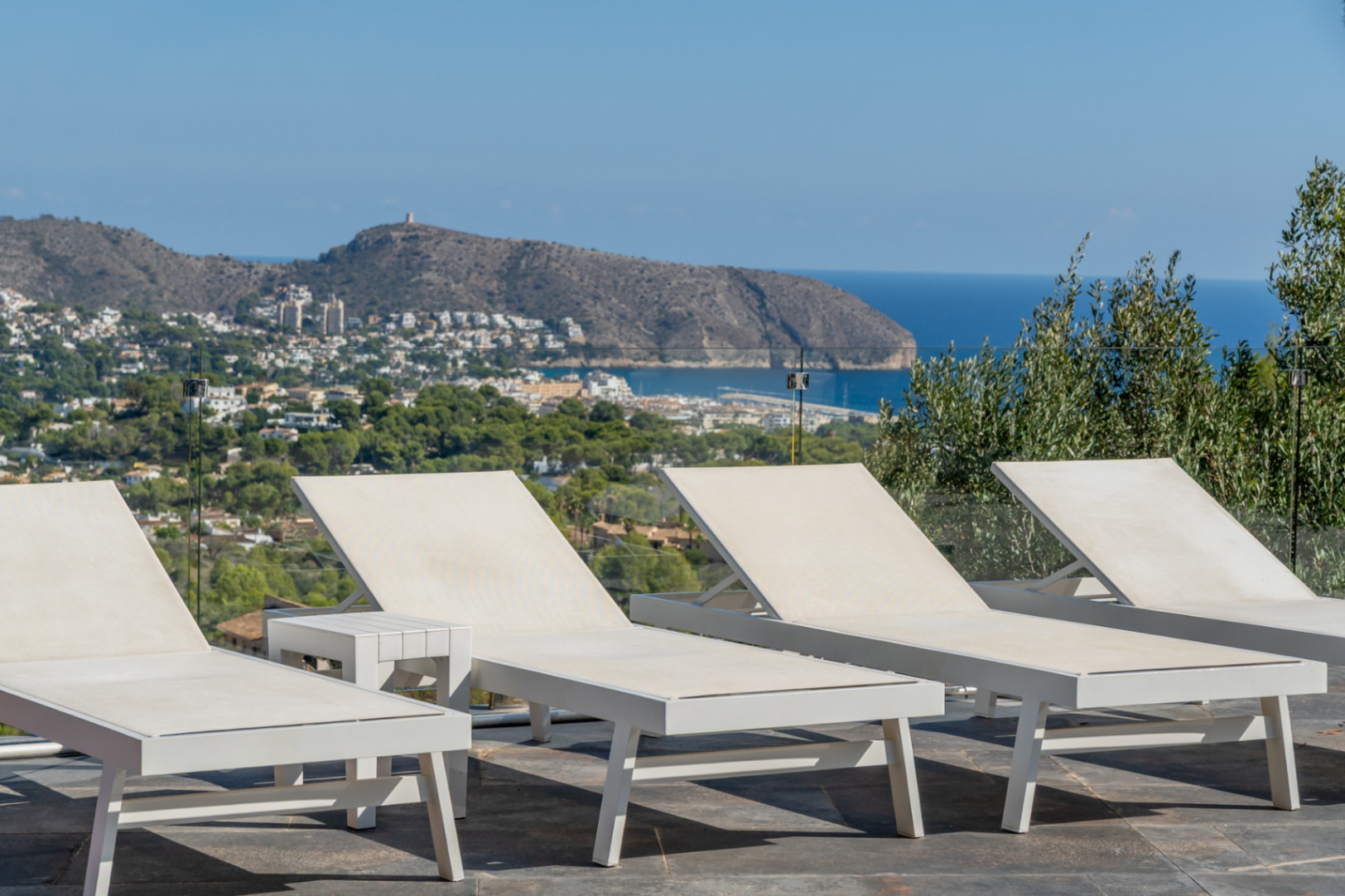 Zona de solárium con tumbonas blancas de diseño y barandilla de cristal, con vistas panorámicas al mar Mediterráneo y al Cap d'Or de Moraira.