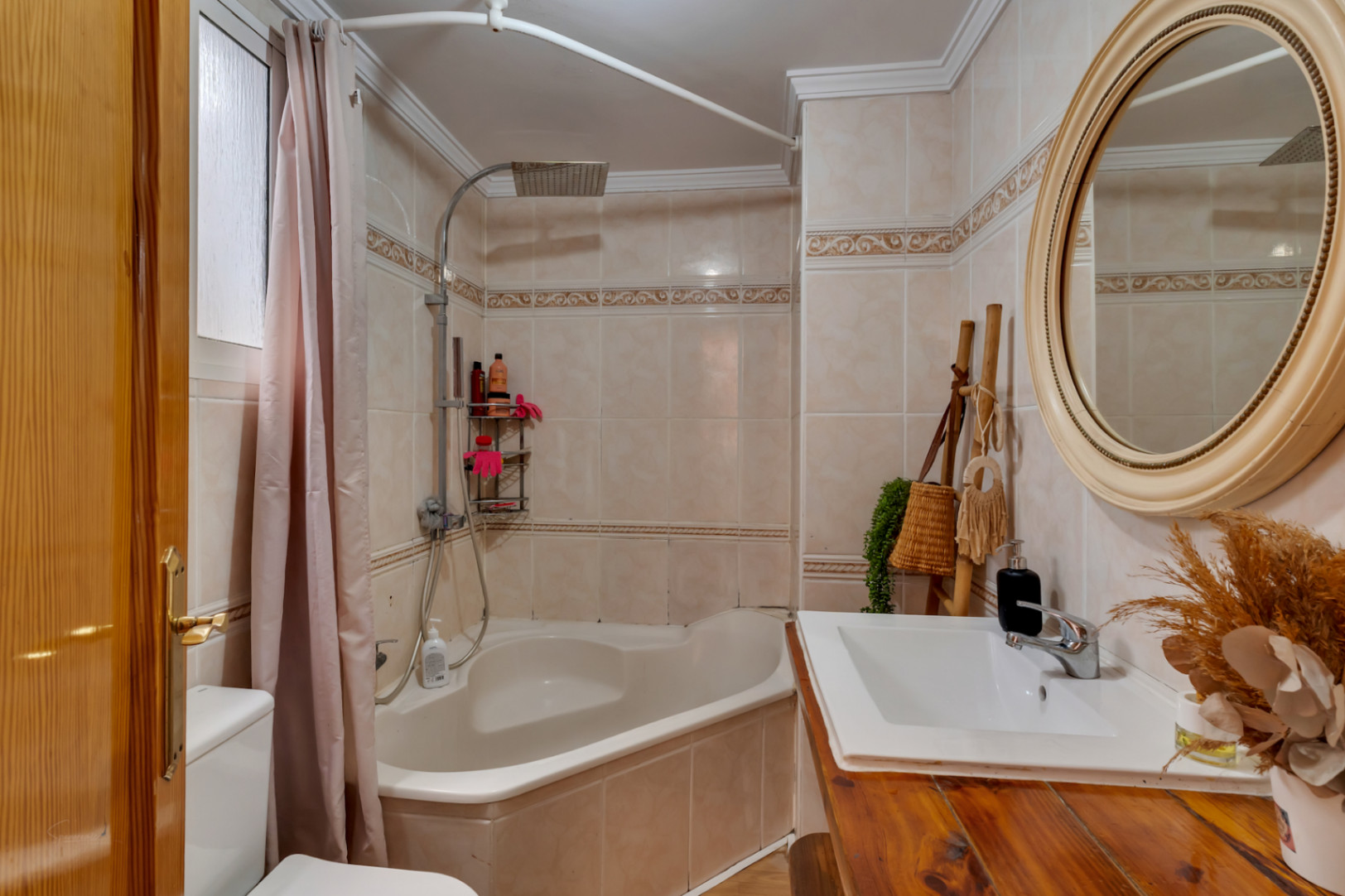 Baño completo con bañera de rincón, encimera de madera rústica y espejo circular sobre azulejos en tonos crema con cenefa decorativa.
