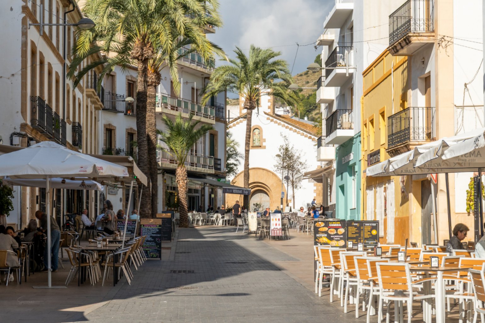 Calle peatonal en el casco antiguo de Jávea con terrazas, palmeras y edificios históricos de fachadas blancas y ocres bajo luz natural.
