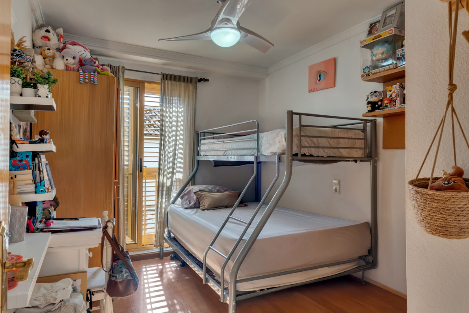 Dormitorio con litera metálica, ventilador de techo y suelos de madera. Incluye acceso directo a un balcón con persianas mallorquinas que aporta abundante luz natural.