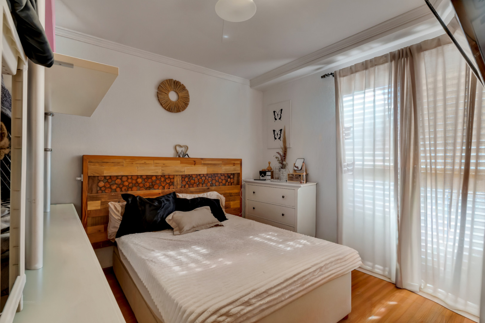 Dormitorio principal con cabecero de madera rústica artesanal, suelos de tarima y ventanal con cortinas que aporta luz natural constante.