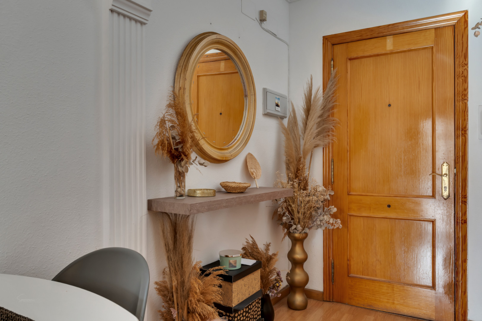 Recibidor con puerta de madera, espejo circular y estante flotante decorado con elementos naturales y tonos cálidos sobre suelo laminado.