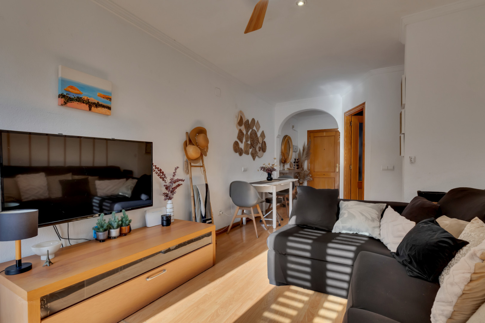 Salón comedor luminoso con suelos de madera, sofá oscuro y mueble de TV. Incluye zona de comedor bajo un arco decorativo y detalles de estilo mediterráneo.