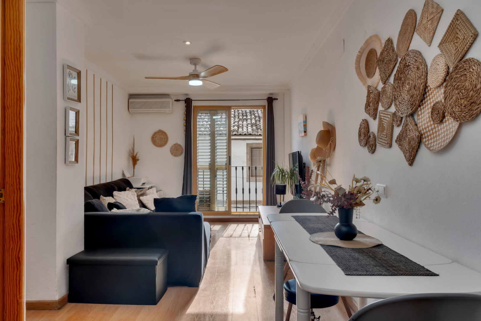 Salón comedor luminoso con suelos de madera, ventilador de techo y acceso directo a un balcón con vistas a los tejados tradicionales del casco antiguo.