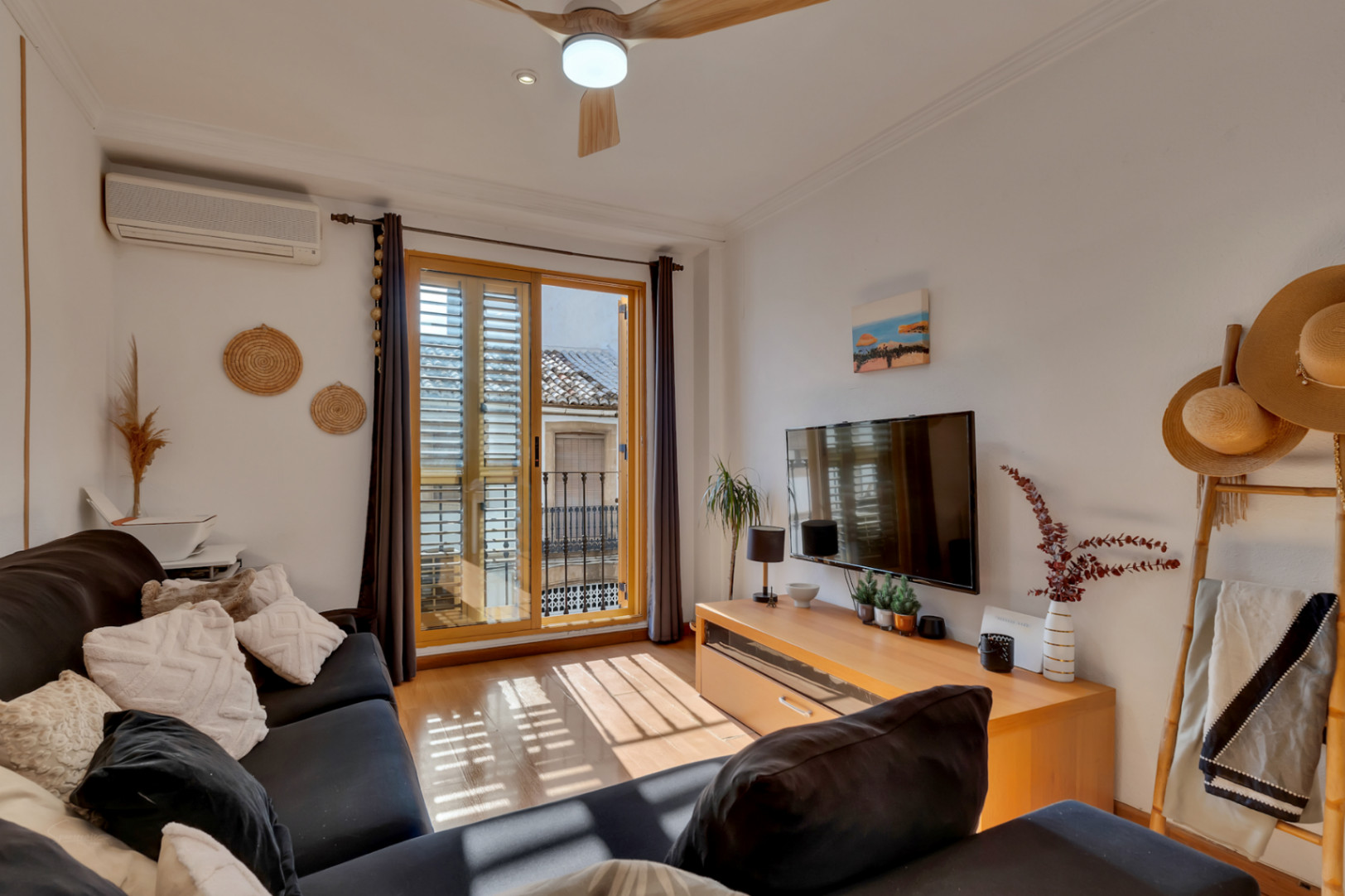 Salón luminoso con suelos de madera, aire acondicionado y balcón con contraventanas tradicionales que ofrece vistas a la arquitectura del casco antiguo.