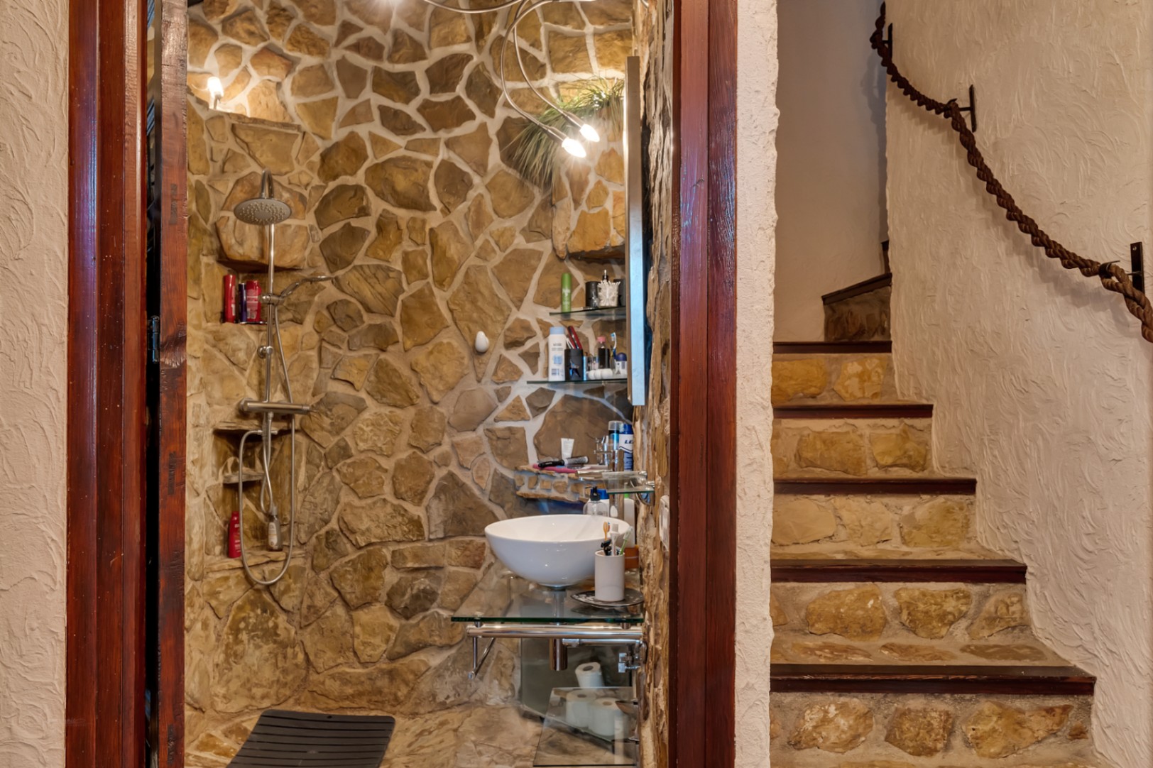 Baño con paredes de piedra natural, lavabo sobre encimera de cristal y ducha integrada. Escalera contigua con peldaños de piedra y pasamanos de cuerda rústica.
