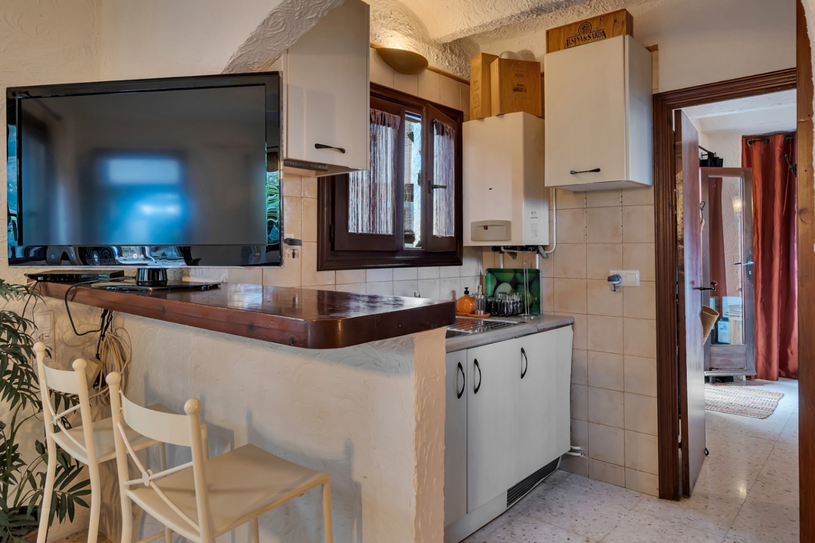 Cocina compacta con barra de madera oscura y taburetes blancos, equipada con armarios altos, fregadero y televisor integrado en el área de desayuno.