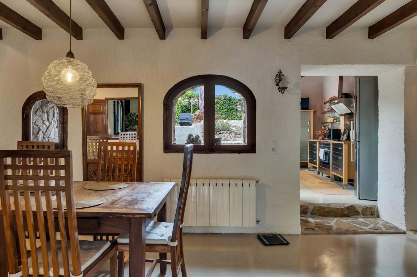 Comedor con vigas de madera, ventana de arco con vistas al jardín y acceso a la cocina moderna a través de un escalón de piedra.