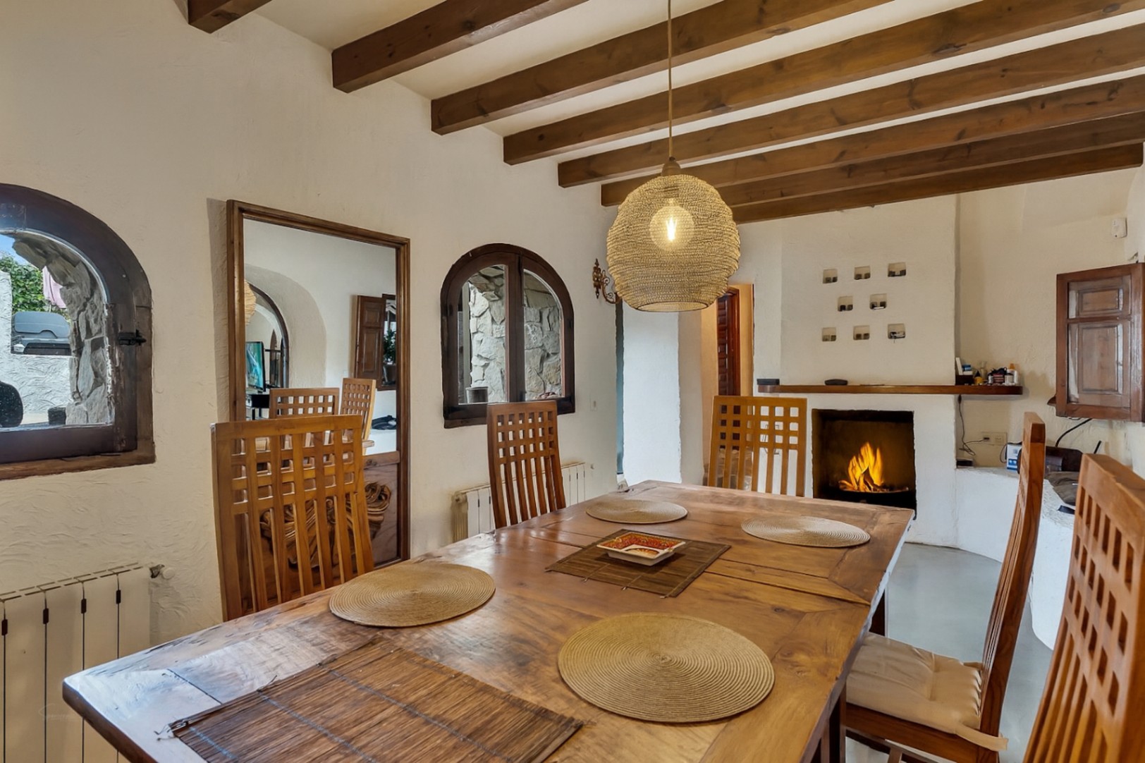 Comedor rústico con vigas de madera vista, mesa de madera maciza y chimenea encendida. Destacan las ventanas de arco y la iluminación cálida mediante una lámpara de fibra natural.