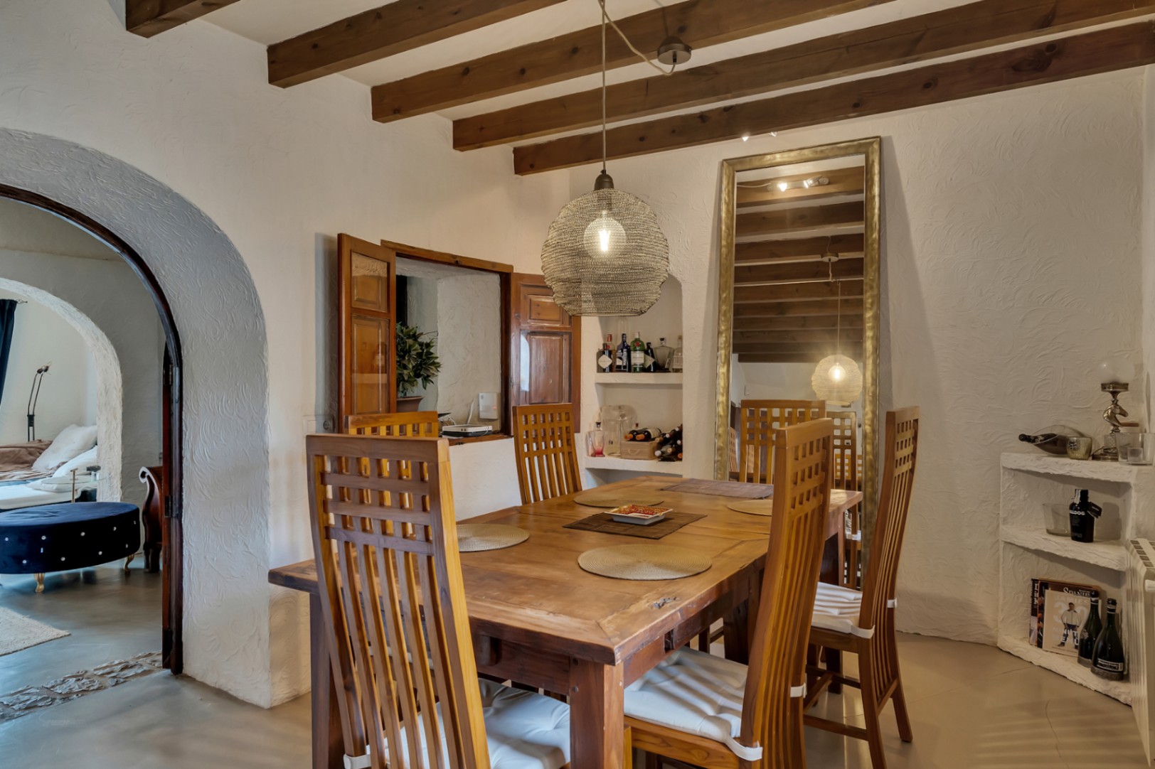 Comedor rústico con vigas de madera vista, mesa de madera maciza y sillas de respaldo alto. Incluye una ventana pasaplatos y un arco que conecta con la estancia contigua.