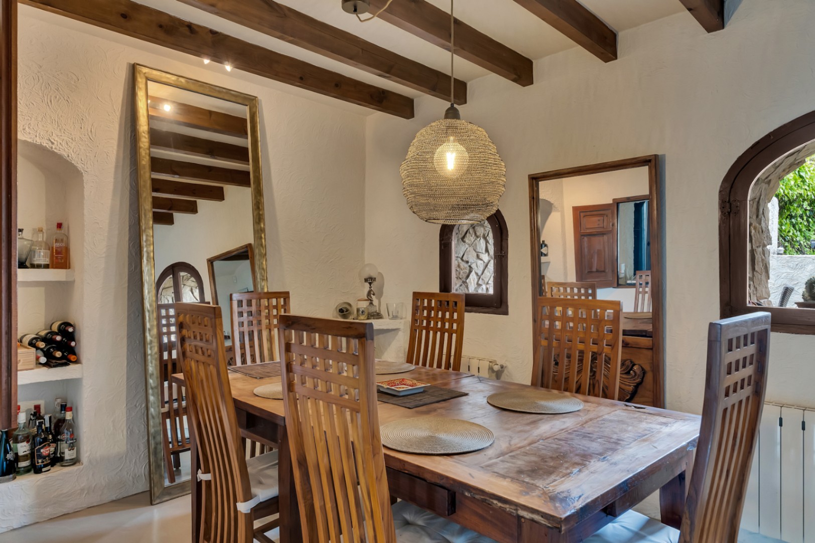 Comedor rústico con vigas de madera vista, mesa maciza y sillas de respaldo alto. Incluye hornacinas integradas, espejos de gran formato y una lámpara de fibra natural.