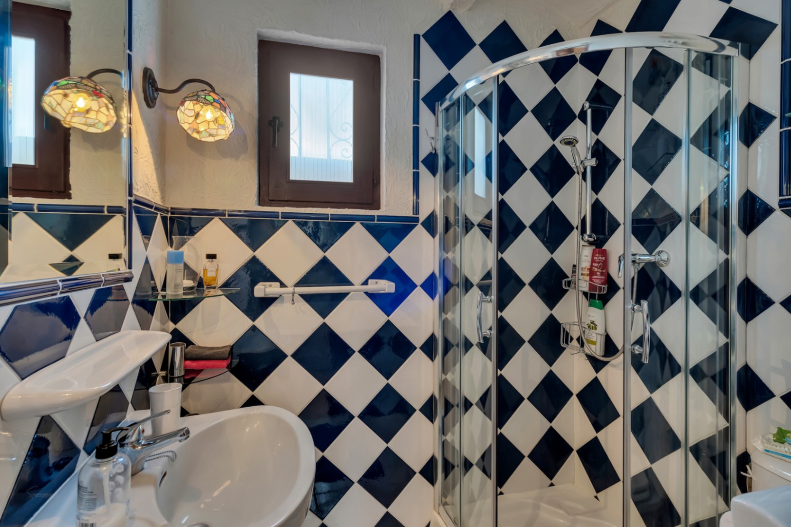Cuarto de baño con azulejos en damero azul y blanco, mampara de ducha curva y apliques decorativos de estilo Tiffany junto a una ventana de madera.