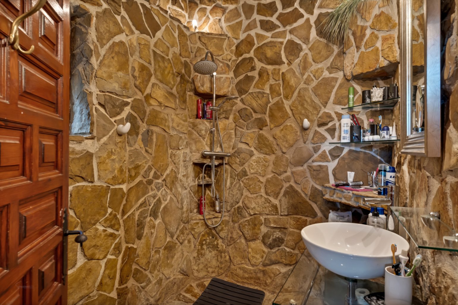 Cuarto de baño rústico con paredes de piedra natural, ducha integrada con grifería de acero y lavabo tipo bol sobre encimera de cristal.