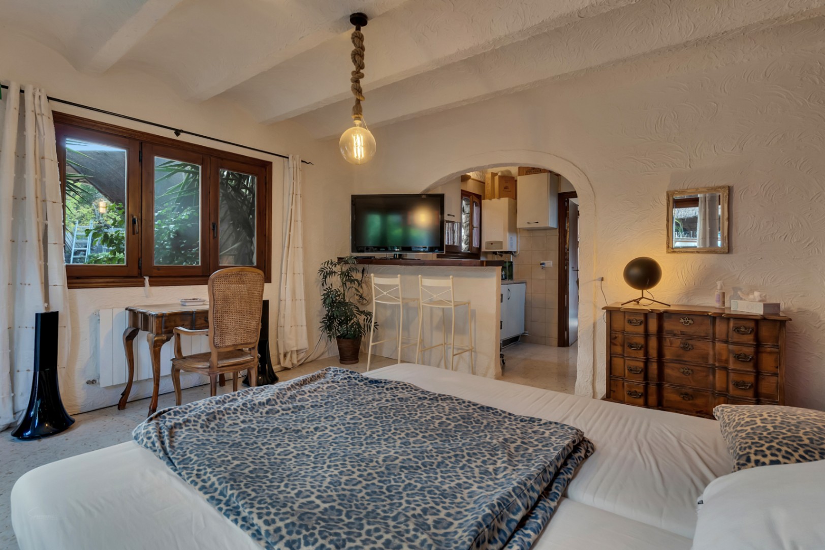 Dormitorio con escritorio de madera, techos con vigas vistas y un arco que comunica con una cocina integrada con barra y taburetes blancos.