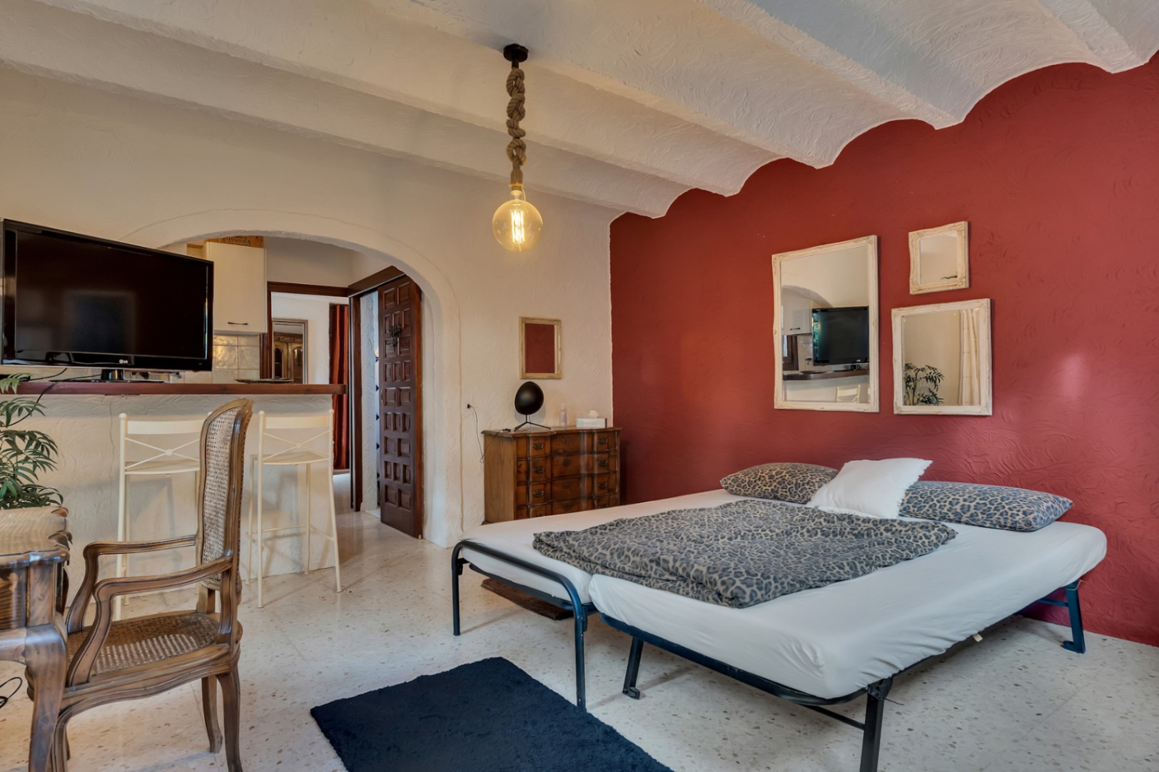 Dormitorio con pared de acento roja, techos abovedados y suelos de terrazo. Incluye un acceso arqueado a la zona de estar y una original lámpara de cuerda.