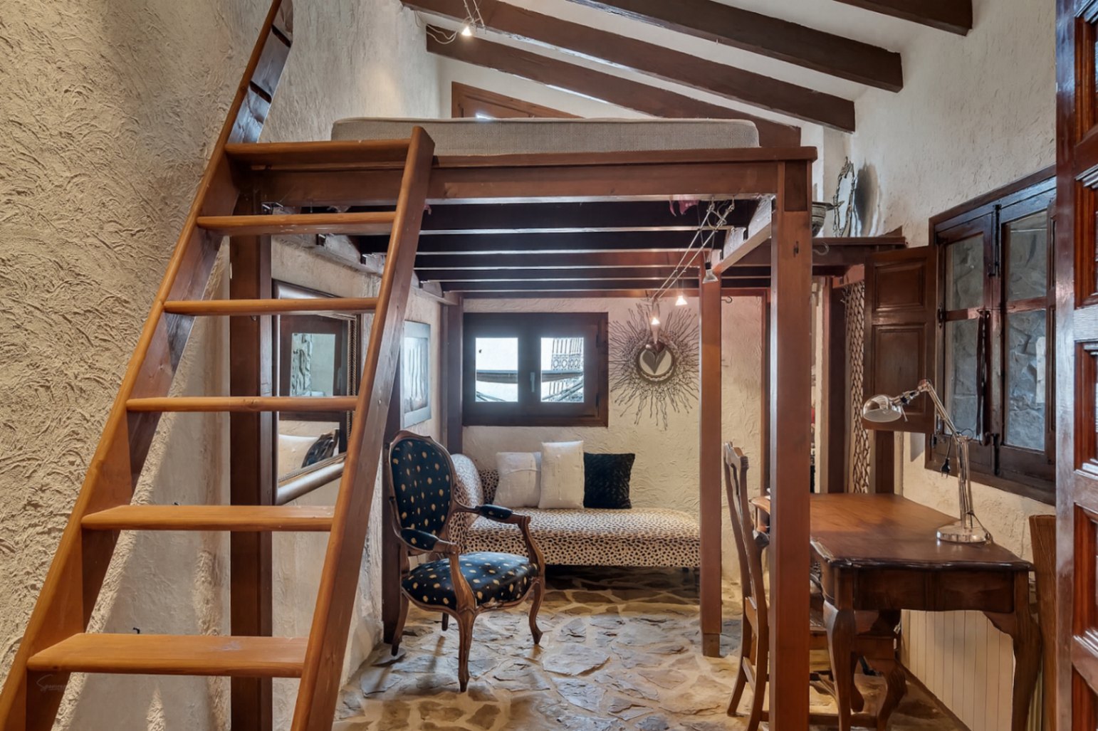 Dormitorio rústico con altillo de madera, vigas vistas y suelo de piedra natural. Incluye una zona de estar bajo la estructura y un escritorio de estilo clásico.