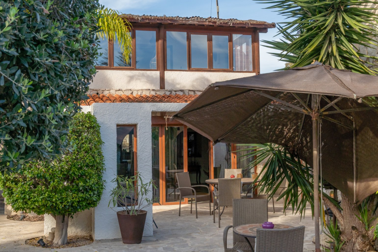 Fachada de la villa con terraza acristalada superior, zona de comedor exterior pavimentada en piedra y vegetación mediterránea madura.