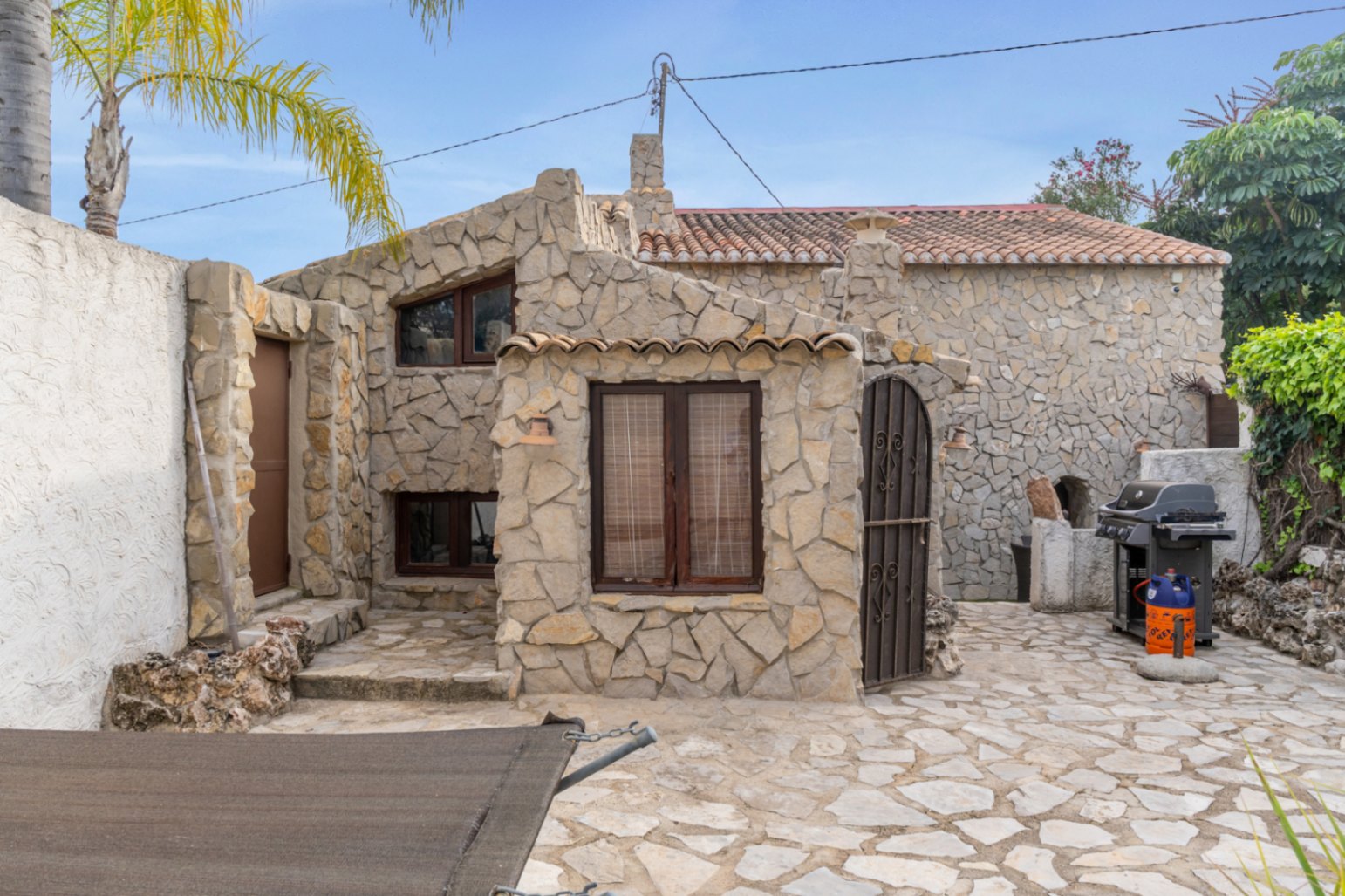 Fachada de piedra natural con carpintería de madera oscura, patio pavimentado y zona de barbacoa exterior junto al apartamento de invitados.