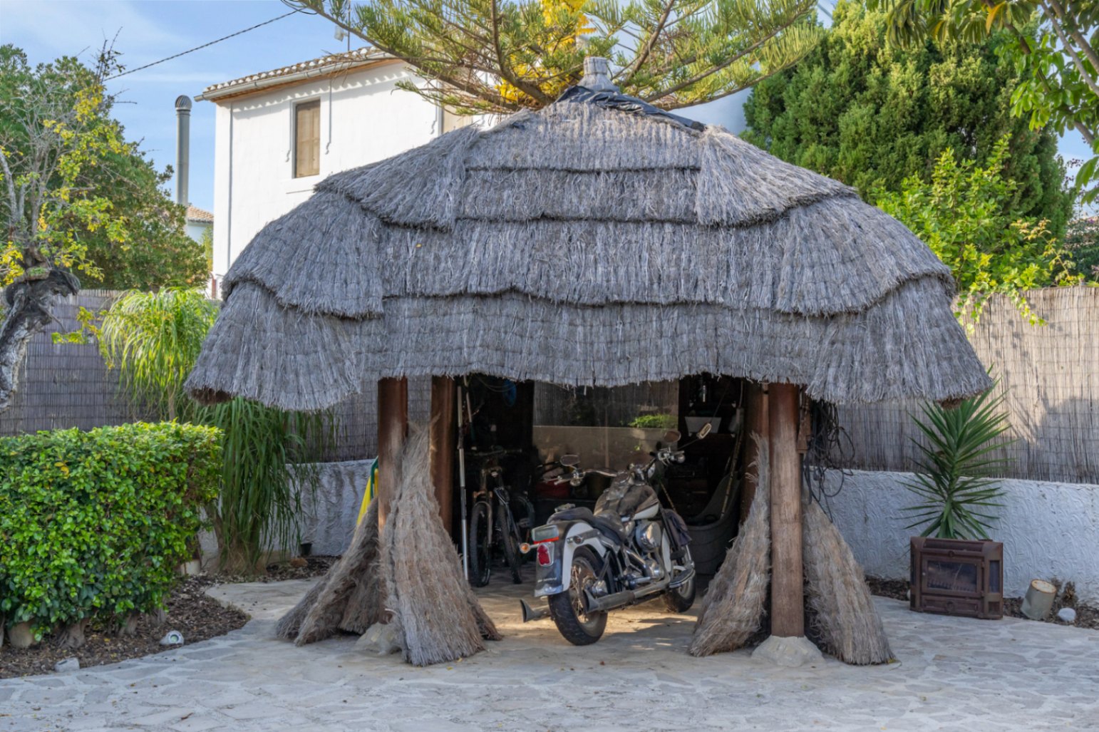 Pérgola de estilo balinés con techo de brezo y vigas de madera, utilizada actualmente como zona de aparcamiento cubierta para motocicletas y bicicletas en el jardín.