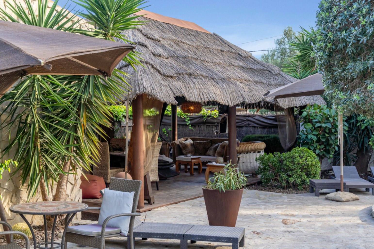 Pérgola de estilo balinés con techo de paja y pilares de madera, situada junto a una terraza de piedra natural con vegetación mediterránea y mobiliario de exterior.
